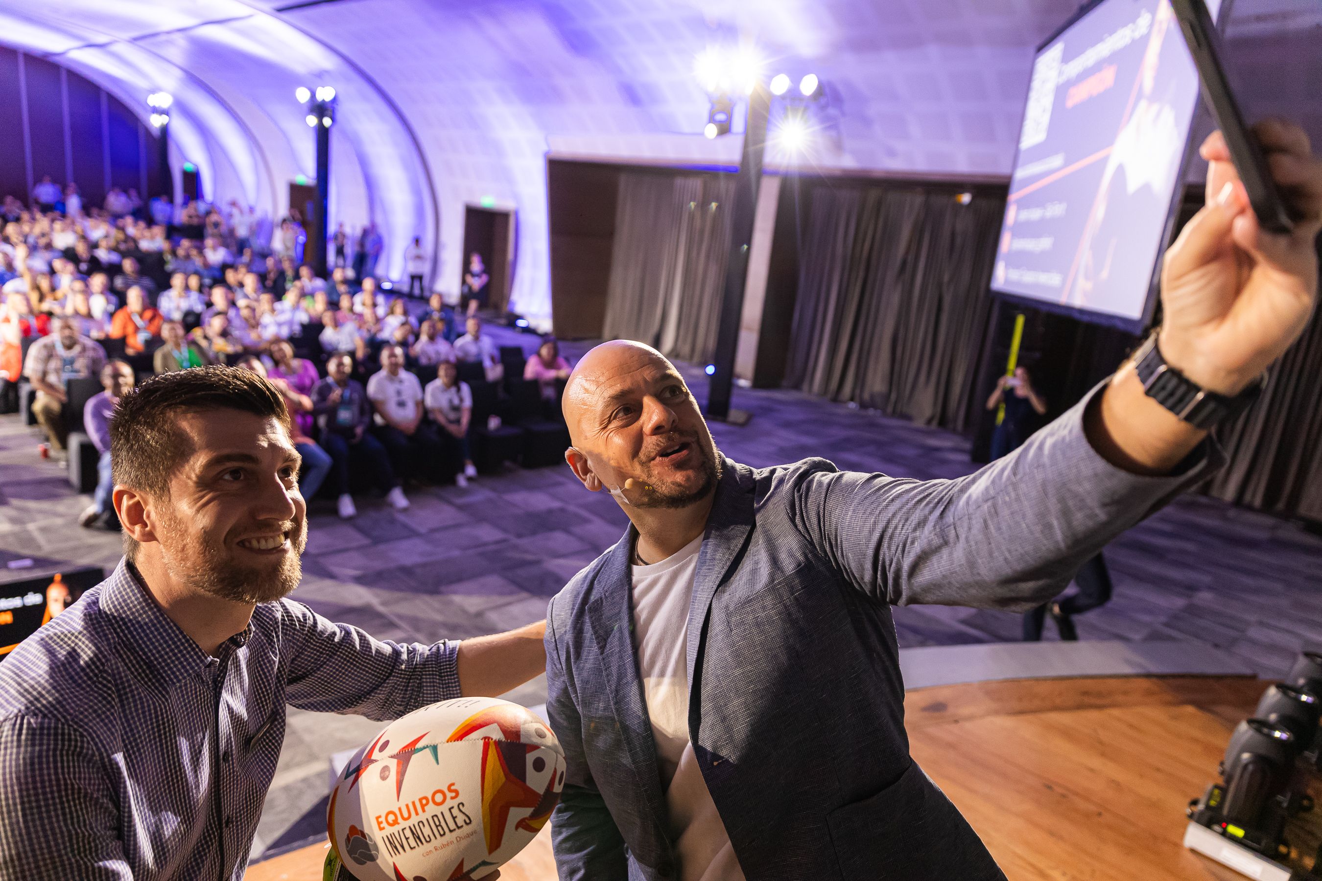 Luiz Bellini and a guest speaker taking a selfie on stage with the audience in the background